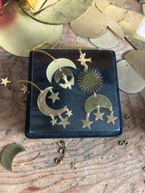 Brass celestial ornaments featuring crescent moons with hanging stars and dove shapes arranged on a dark square base against a wooden surface with scattered brass pieces.