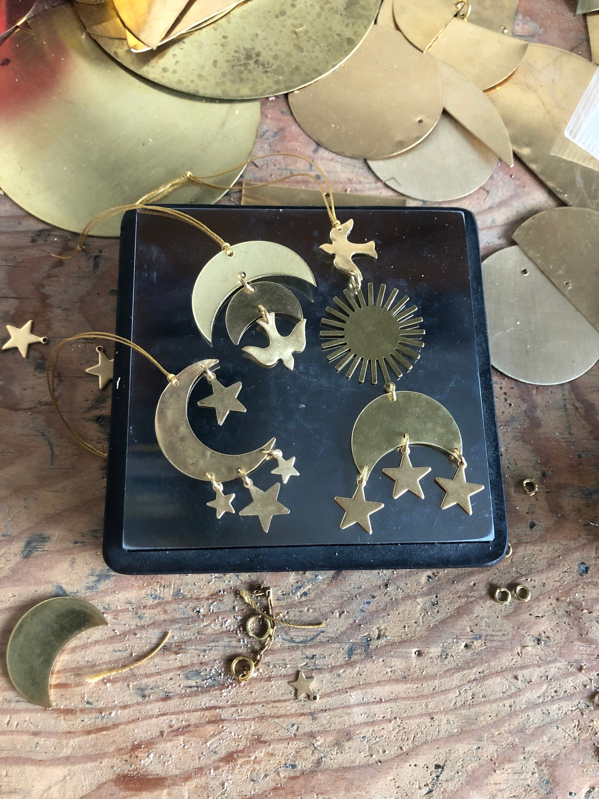 Brass celestial ornaments featuring crescent moons with hanging stars and dove shapes arranged on a dark square base against a wooden surface with scattered brass pieces.