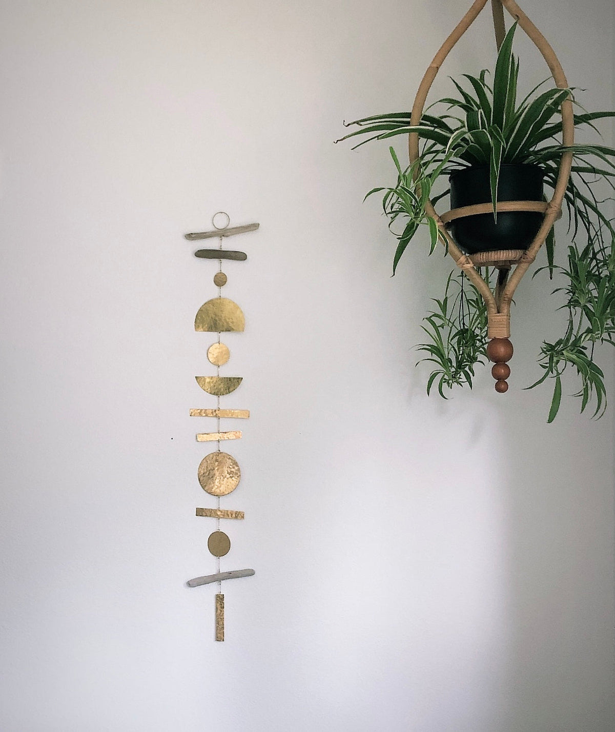 Vertical hanging mobile with gold and silver geometric shapes against a pale wall, next to a wooden hanging planter holding green plants.