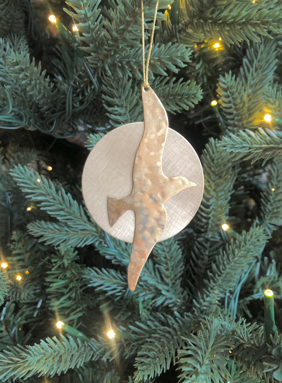 Hammered copper bird silhouette ornament suspended in front of a smooth, circular silver disc on a green pine tree with small warm lights.