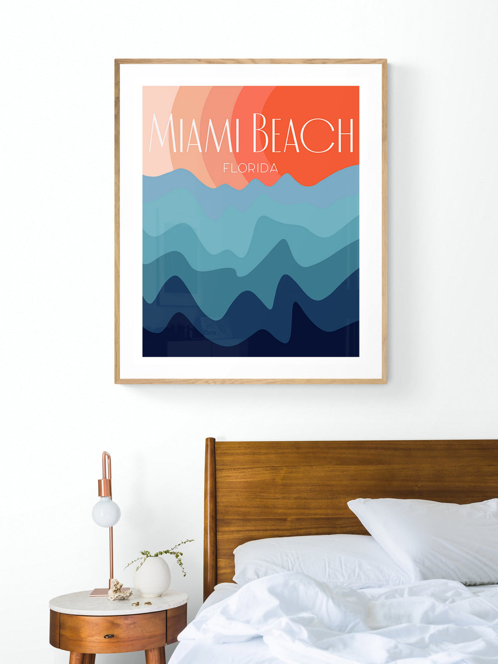 Modern art print with abstract blue waves and orange sky background, titled 'Miami Beach Florida,' framed and hung above a wooden bed in a minimalist bedroom.