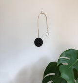 Minimalist wall suncatcher with a black circular base and a curved gold metal rod ending in a clear teardrop crystal, hanging above green monstera leaves.
