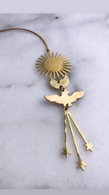 Brass ornament featuring a sunburst disc, a smaller round disc, and a bird shape with three star-tipped hanging strands on a leather cord, photographed on light marble.