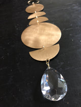 Gold-toned metal discs in varying sizes linked vertically, ending with a large faceted clear teardrop crystal against a dark background.