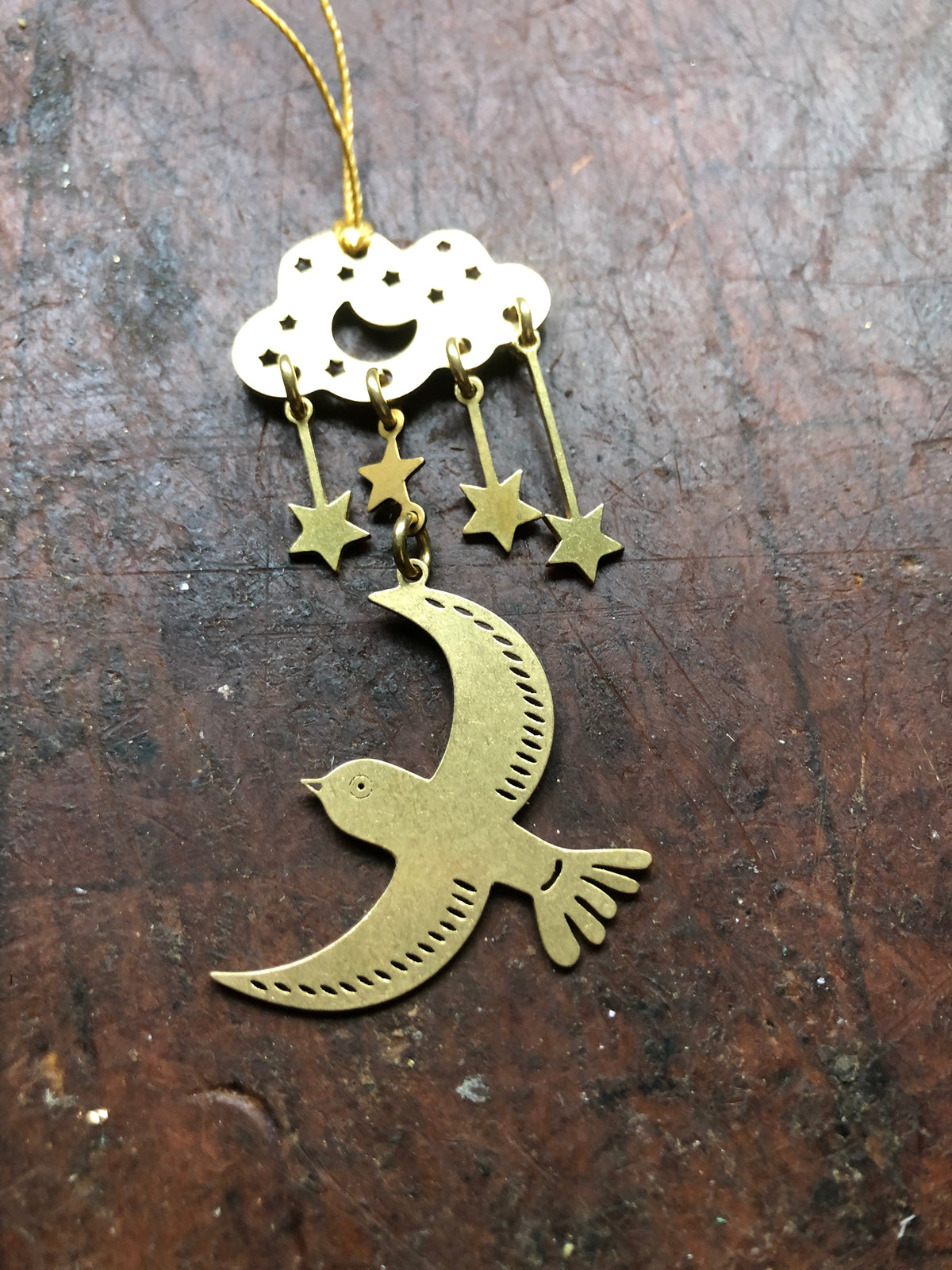 Brass ornament featuring a cloud with three dangling stars above a bird with engraved feather details, all suspended on a dark textured surface.