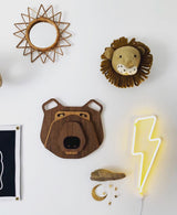 Wall display with a wooden geometric sunburst mirror, plush lion head with fabric mane, layered wooden bear face sculpture, and a lit yellow neon lightning bolt on white wall.