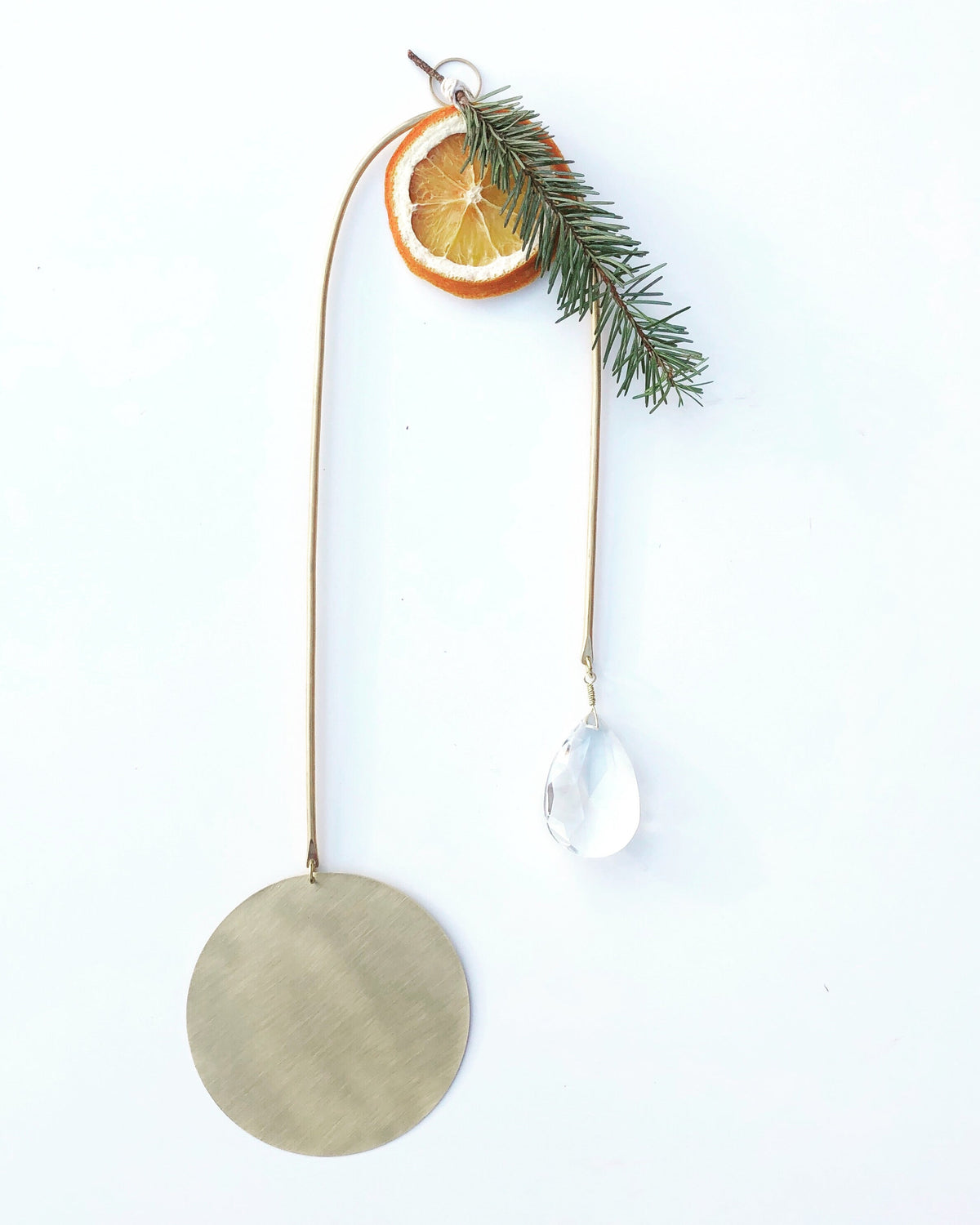 Minimal suncatcher with a large brushed gold disc, hanging clear teardrop crystal, dried orange slice, and a small green pine sprig on thin wire.