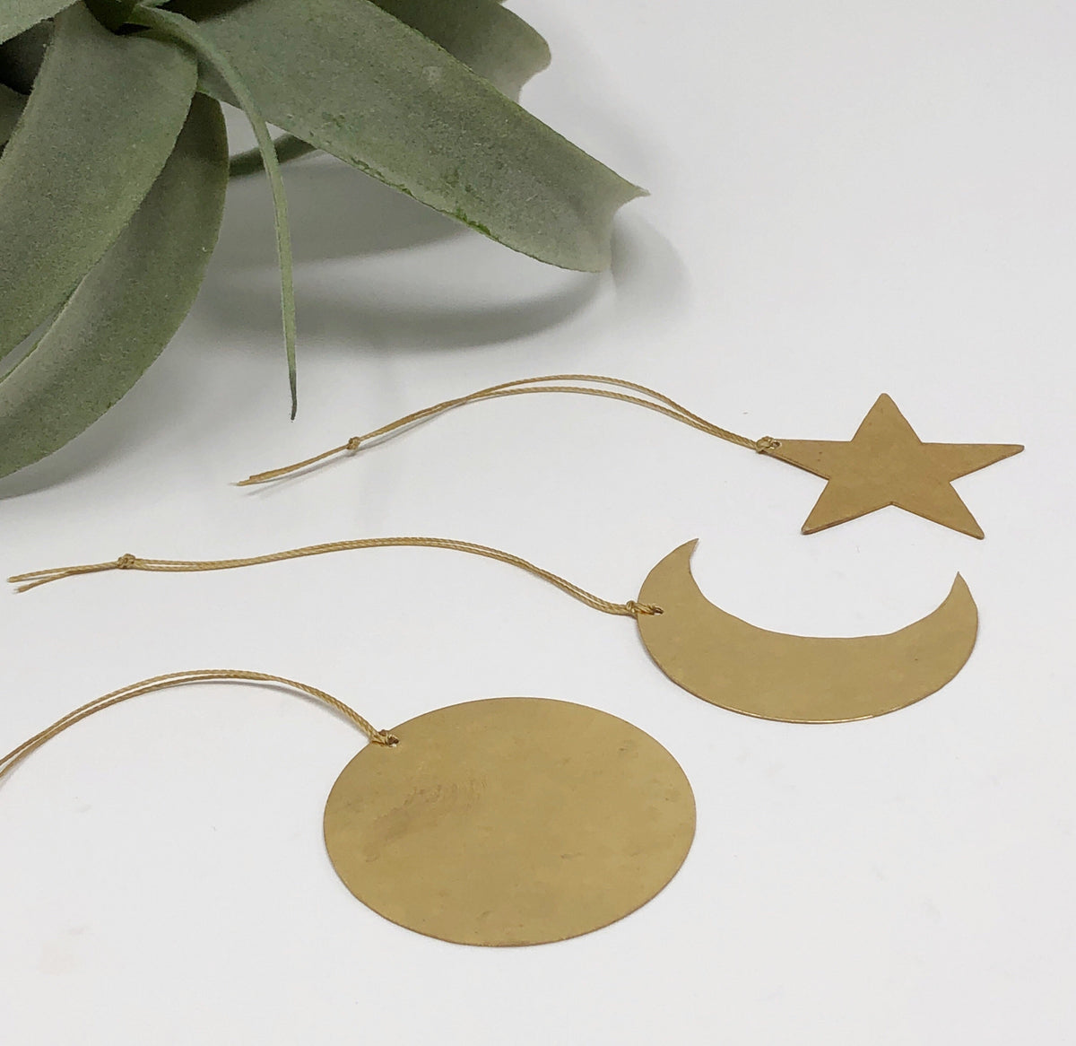 Three brass celestial ornaments shaped as a star, crescent moon, and circle, each hanging on thin gold cords against a white background with green foliage nearby.