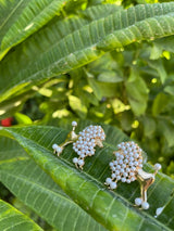 Pair of gold-tone poodle-shaped earrings adorned with clustered white pearls, resting on vibrant green long leaves with lush foliage background.