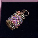 Small gold-tone cylindrical charm encrusted with iridescent Swarovski crystals, featuring a keyring attachment, displayed on a dark velvet background.