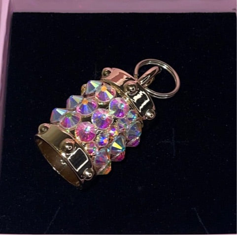 Small gold-tone cylindrical charm encrusted with iridescent Swarovski crystals, featuring a keyring attachment, displayed on a dark velvet background.