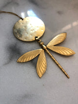 Golden dragonfly ornament with detailed veined wings attached to a round hammered metal tag on a gray marble surface.