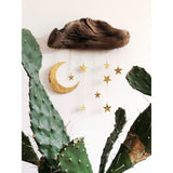 Wall sculpture with textured brown wood piece and hanging gold crescent moon and stars, displayed above green cactus leaves against a white wall.