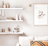 Minimalist living room corner with white floating shelves holding ceramic vases and wooden decor, and a white couch with a patterned brown and black pillow beneath a framed botanical print.