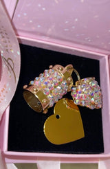 Two gold-tone charms encrusted with iridescent Swarovski crystals shaped like a cylinder and a heart, paired with a smooth gold heart pendant inside a pink box.
