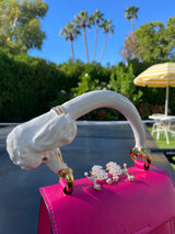 White sculpted poodle-shaped bag handle and matching pearl poodle earrings displayed on a bright pink handbag with a sunny garden background.