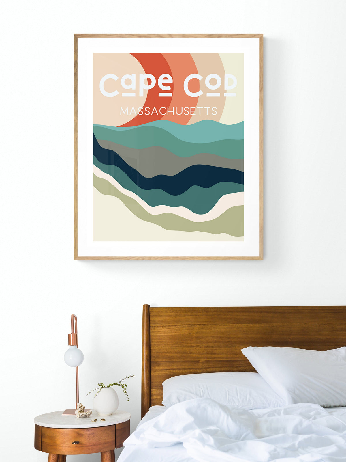 Abstract Cape Cod map print with wavy, layered colors in teal, navy, red, and beige, framed and hanging above a wooden bed and round bedside table.