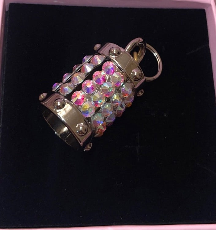 Silver cylindrical charm decorated with rows of iridescent Swarovski crystals, featuring metal caps with rivet details and a keyring attachment, displayed on black fabric.