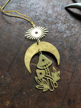Brass ornament with a sunburst, crescent moon, and two stylized mushrooms with star and crescent cutouts, set on a textured dark surface.