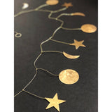 Hammered brass garland with celestial shapes including stars, moons, and circles linked by delicate chain on a dark background.