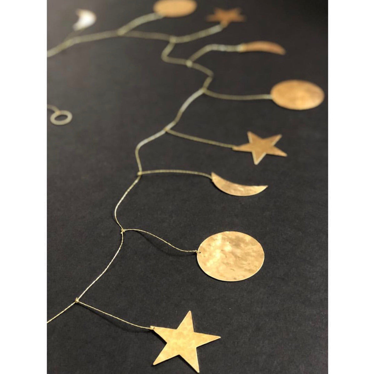 Hammered brass garland with celestial shapes including stars, moons, and circles linked by delicate chain on a dark background.