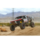 Black and red off-road race truck with Nexgen and General Tire logos airborne over desert terrain with mountains and power lines in the background