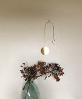 Minimalist wall suncatcher with a thin gold wire, a round gold disk, and a hanging clear crystal teardrop above dried brown foliage in a blue-green glass vase against a white wall.