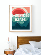 Modern framed print above bed showing abstract sun and layered waves in coral, teal, and dark blue with text 'Mackinac Island Michigan'.
