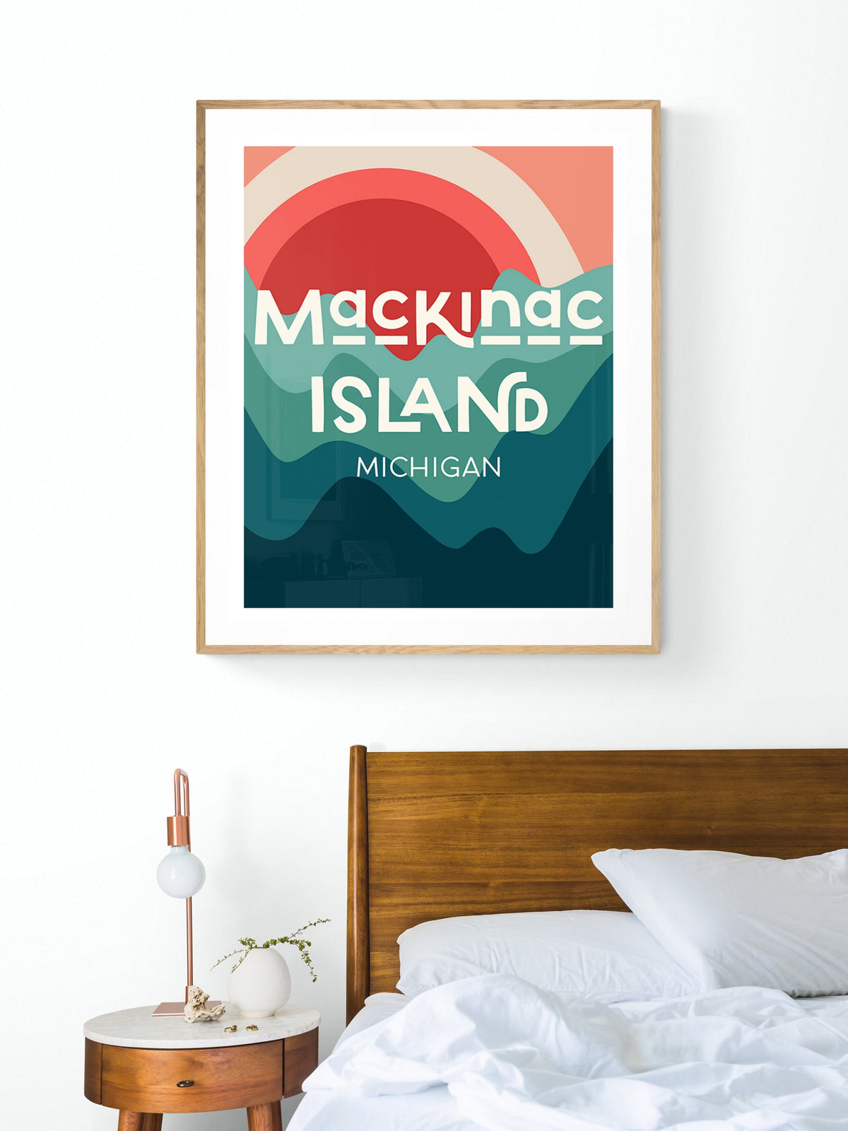 Modern framed print above bed showing abstract sun and layered waves in coral, teal, and dark blue with text 'Mackinac Island Michigan'.