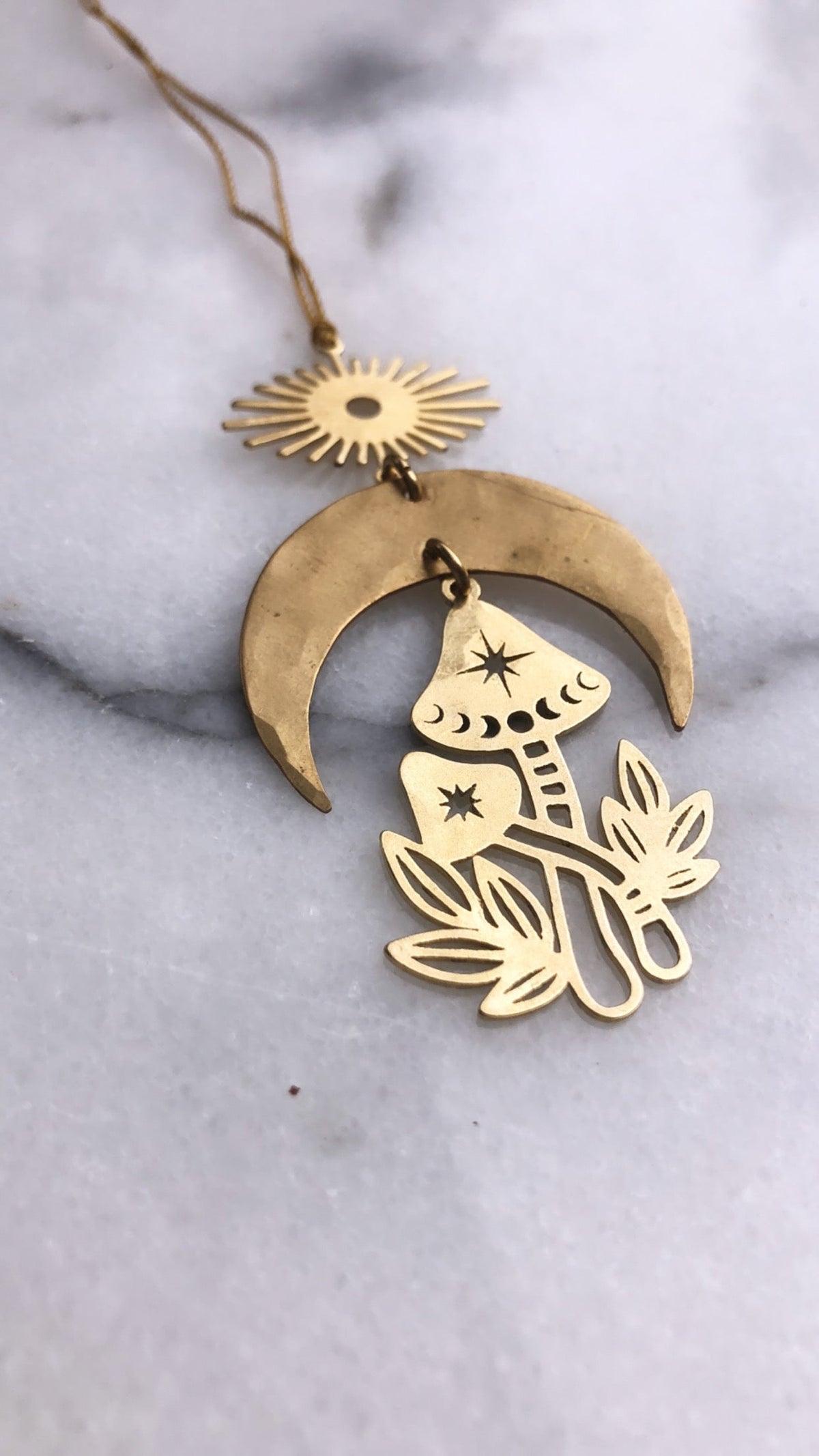 Gold-toned necklace pendant featuring a crescent moon, sunburst, and stylized mushroom with star and dot details on a soft marble surface.