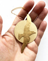 Hand holding a round beige ornament featuring a gold-toned metal silhouette of a bird in flight with a thin gold hanging string.