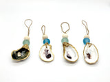 Four sea glass ornaments each featuring a painted oyster shell with gold trim, a round wooden bead, and a colored sea glass bead in shades of blue and green, hanging on twine loops.
