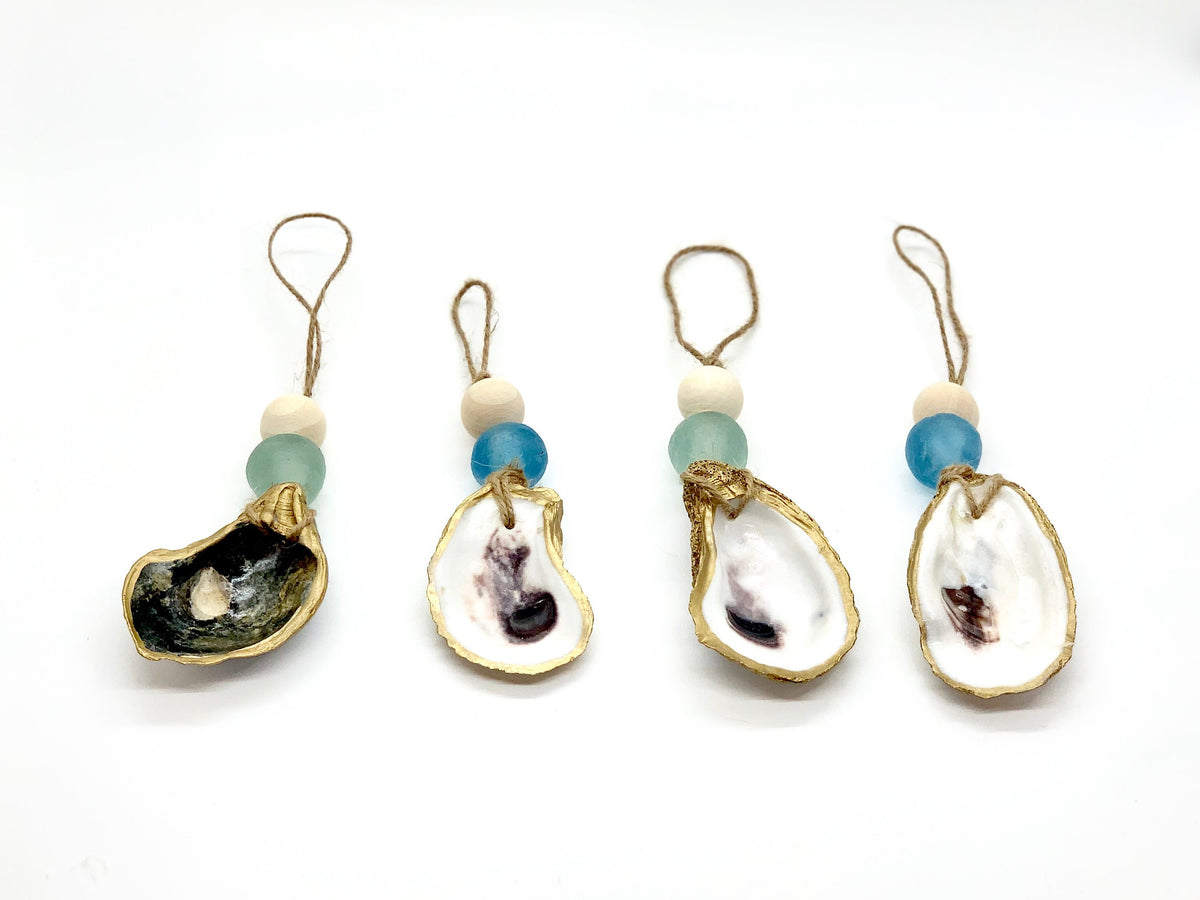 Four sea glass ornaments each featuring a painted oyster shell with gold trim, a round wooden bead, and a colored sea glass bead in shades of blue and green, hanging on twine loops.