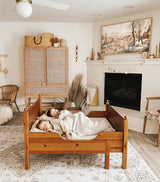 Two children sleeping under a cream knitted blanket on a wooden bed in a beige and white living room with a fireplace, woven chairs, and a rustic cabinet.