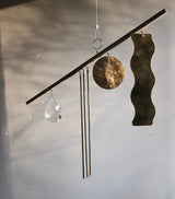 Minimalist mobile with a horizontal wooden rod suspending a teardrop-shaped clear crystal, three thin metal rods, a hammered round brass disc, and a wavy brass strip against a soft gray background.