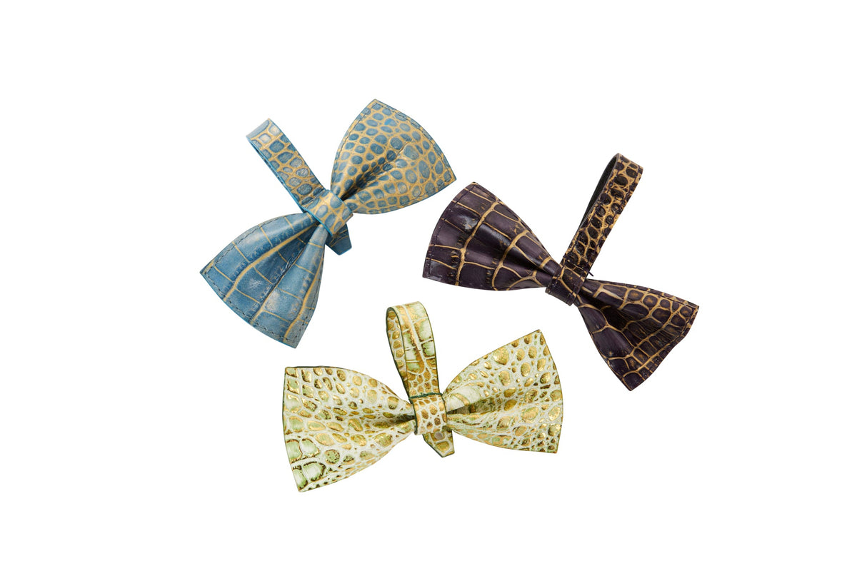 Set of three Italian leather bowties embossed with crocodile patterns in blue, dark purple with gold highlights, and greenish-gold tones, arranged on a white background.