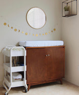 Brass garland of hanging celestial shapes including moons and stars strung beneath a round mirror with wood frame in a nursery corner over a wooden changing table and white rolling cart.