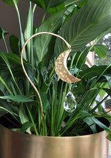 Hammered gold crescent moon hanging from a curved gold metal stick with a faceted teardrop crystal, placed in a green leafy plant in a gold planter near a window.
