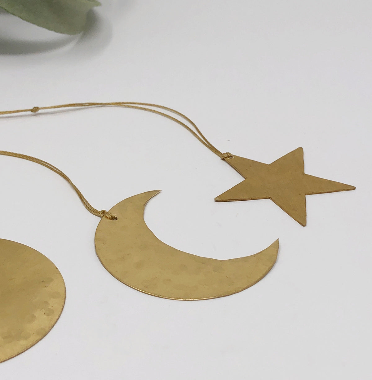 Three brass celestial ornaments including a circle, crescent moon, and star, each hanging from thin brown cords against a white background.