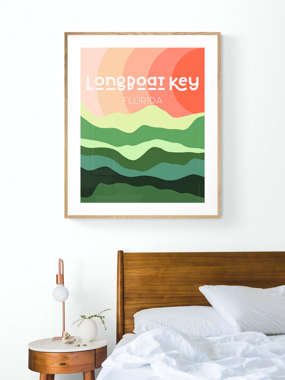 Modern art print titled 'Longboat Key Florida' with layered abstract green hills and orange sun, framed and hung above a wooden bed and round bedside table.