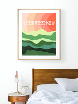 Modern art print titled 'Longboat Key Florida' with layered abstract green hills and orange sun, framed and hung above a wooden bed and round bedside table.