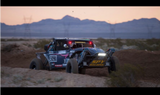Off-road racing buggy with number 5090 equipped with bright front LED lights, driving on dirt terrain during sunset with mountains and power lines in background.