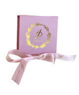 Light pink square box with shiny gold wreath and stylized initial A, accompanied by matching pink satin ribbon printed with gold script and initials.