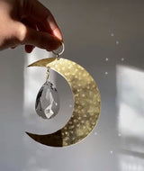 Hand holding a crescent moon-shaped metallic suncatcher with a large faceted crystal teardrop hanging inside, casting light reflections on a neutral background.