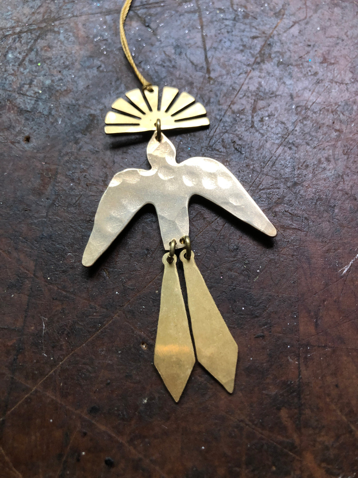 Hammered metal bird ornament with two dangling geometric tail pieces hanging beneath a semicircular top piece with radiating cutouts, set against a dark textured background.