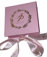 Light pink square gift box with gold laurel wreath and script logo on top, tied with matching pink satin ribbon printed with gold text and logo.