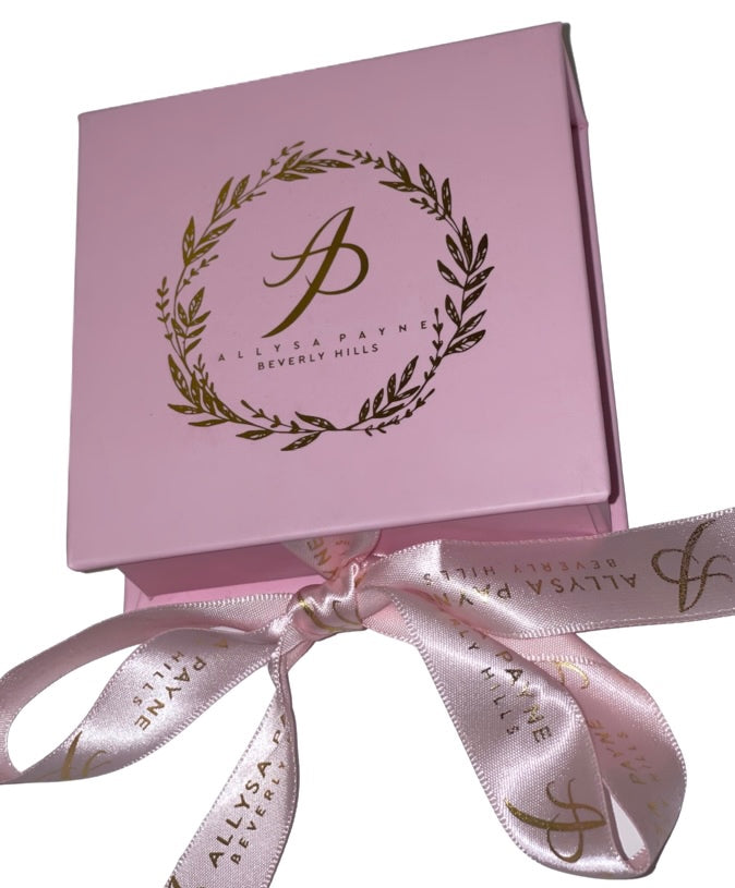 Light pink square gift box with gold laurel wreath and script logo on top, tied with matching pink satin ribbon printed with gold text and logo.