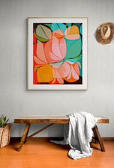 Framed abstract botanical print featuring overlapping shapes in orange, pink, teal, and green on a dark background, displayed above a wooden bench with a white cloth.
