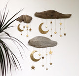 Three driftwood pieces mounted on a white wall with dangling hammered gold-tone crescent moons and stars, arranged in a descending cluster beside green plant leaves.