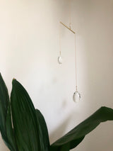 Delicate hanging suncatcher with two faceted crystal pendants suspended from thin gold chains attached to a horizontal gold bar, against a light wall and large green leaves below.